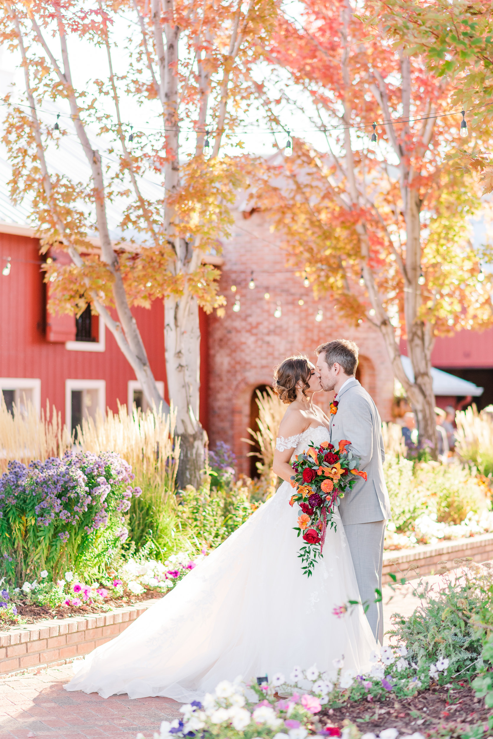 Bride and groom kiss in front of a red barn surrounded by trees with red and yellow leaves.