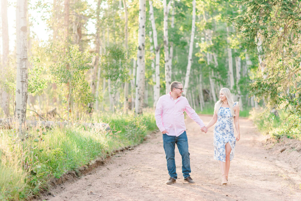 Man and woman walking hand in hand down a wooded trail during their outdoor engagement session in Colorado.