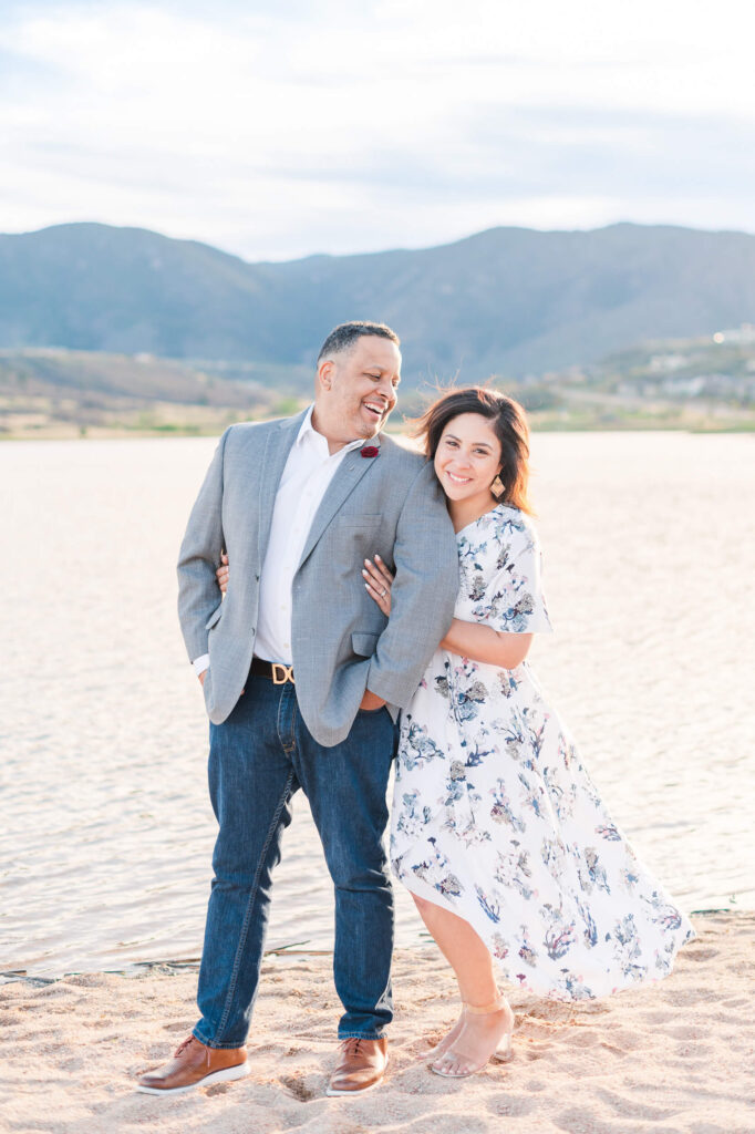 A couple poses on a sandy beach in front of a lake for their engagement photos, with a view of mountains in the background.