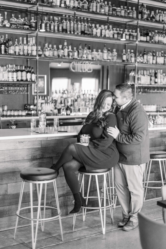 A woman sits at a bar top holding a drink while her fiance stands behind her during their engagement session in Colorado Springs.