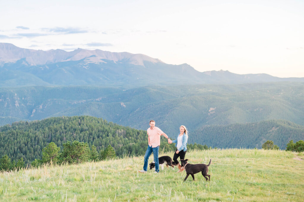 A couple walks hand in hand on a green grassy hill overlooking Pikes Peak with their two dogs.
