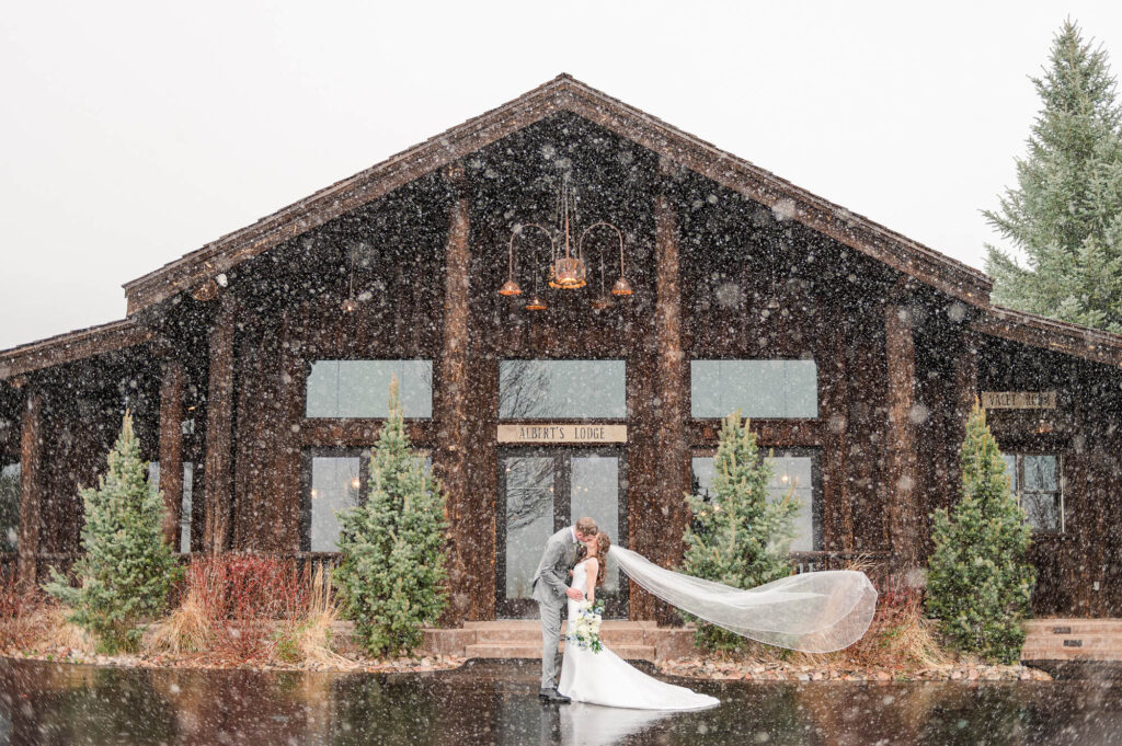 A bride and groom kiss in the snow in front of Spruce Mountain Ranch in Larkspur, Colorado.