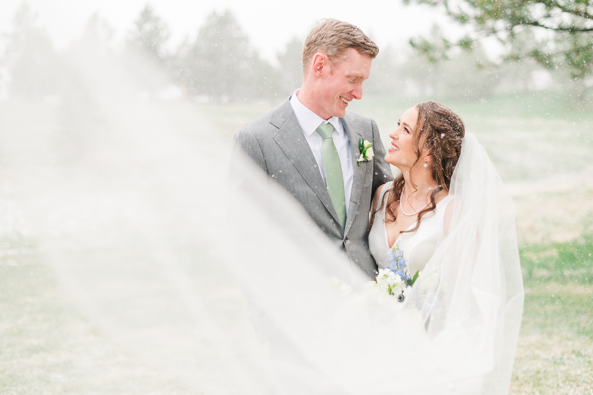 A bride's long veil comes toward us as she looks at her groom outside on their snowy wedding day.