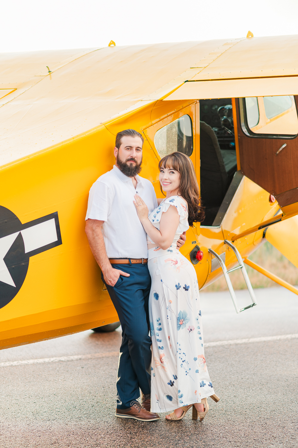 Man and woman pose leaning against a yellow vintage airplane during engagement photos.