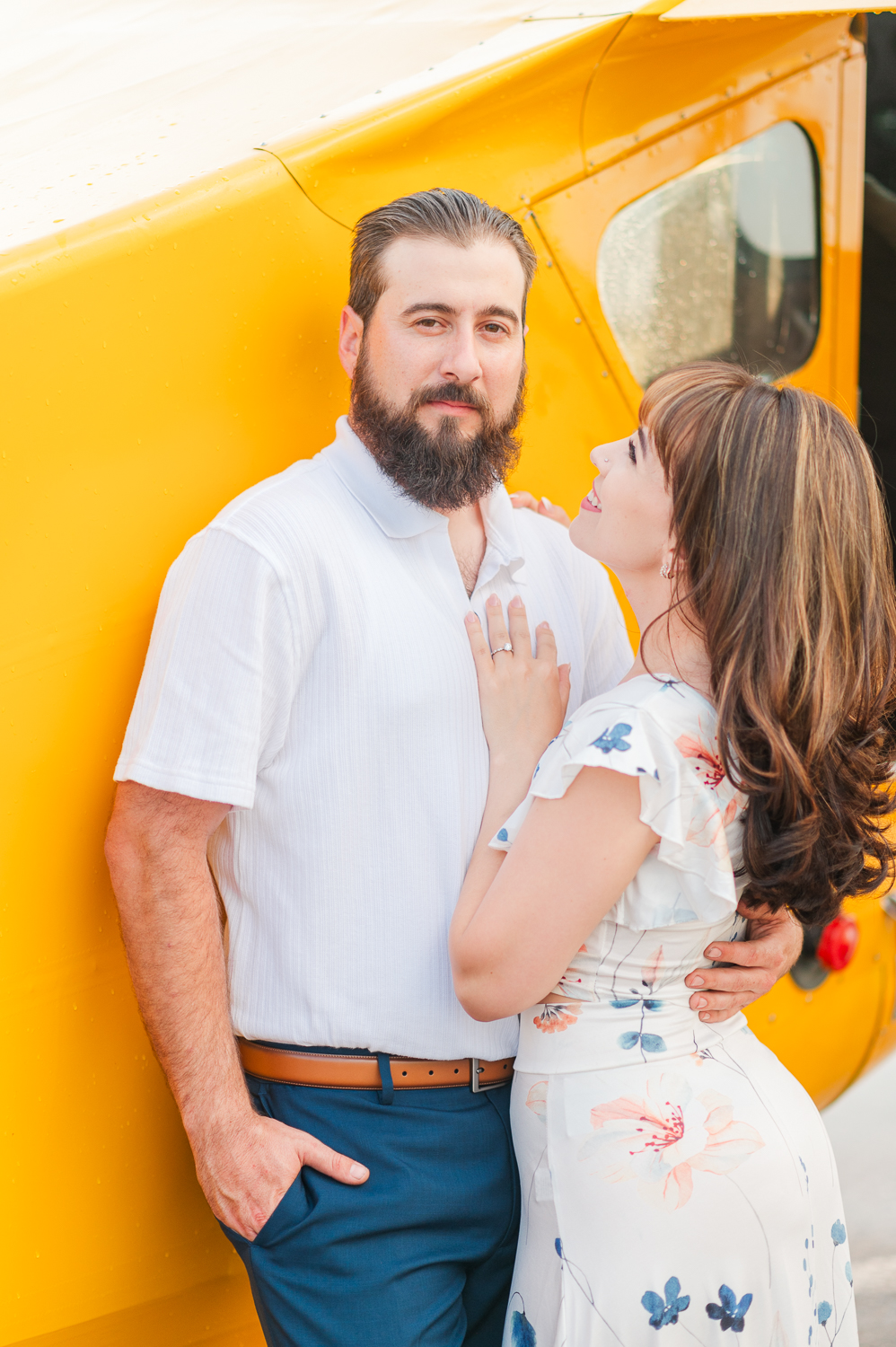 A man looks directly at the camera while leaning against a yellow airplane as his fiance smiles up at him during their engagement session.